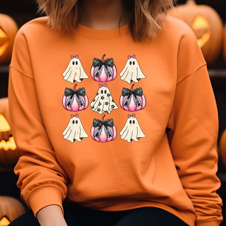 May include: Orange sweatshirt featuring a grid pattern of Halloween-themed graphics. The design includes pink pumpkins with black bows and ghosts with bows. The pumpkins have a lace trim. The ghosts are white with black eyes.