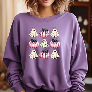 May include: A purple sweatshirt featuring a Halloween-themed design. The design includes cartoon ghosts and pink pumpkins with black bows. The ghosts are white with black eyes, and the pumpkins have black polka dots. The sweatshirt is a solid purple color.