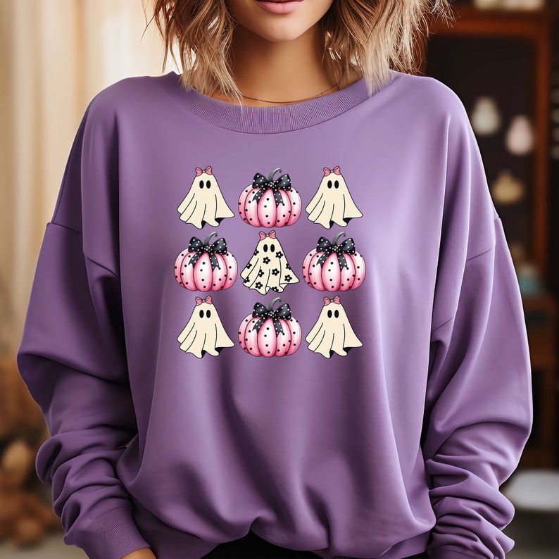 May include: A purple sweatshirt featuring a Halloween-themed design. The design includes cartoon ghosts and pink pumpkins with black bows. The ghosts are white with black eyes, and the pumpkins have black polka dots. The sweatshirt is a solid purple color.