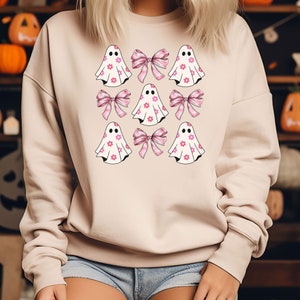 May include: Beige sweatshirt featuring a pattern of ghost figures with pink flower details and pink bows. The ghosts have black eyes and are decorated with small pink flowers. The bows are a light pink color.