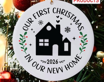 May include: A round white ornament featuring a black silhouette of a house in the center with the text "OUR FIRST CHRISTMAS IN OUR NEW HOME" and "2026" printed in black. The design includes green sprigs with red berries and small snowflakes.