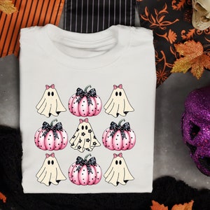 May include: White t-shirt featuring a Halloween design with ghosts and pumpkins. The design includes pink pumpkins with black bows and polka dots, and ghosts with pink bows. A fun and festive shirt for the spooky season.
