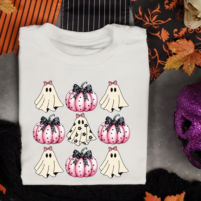 May include: White t-shirt featuring a Halloween design with ghosts and pumpkins. The design includes pink pumpkins with black bows and polka dots, and ghosts with pink bows. A fun and festive shirt for the spooky season.