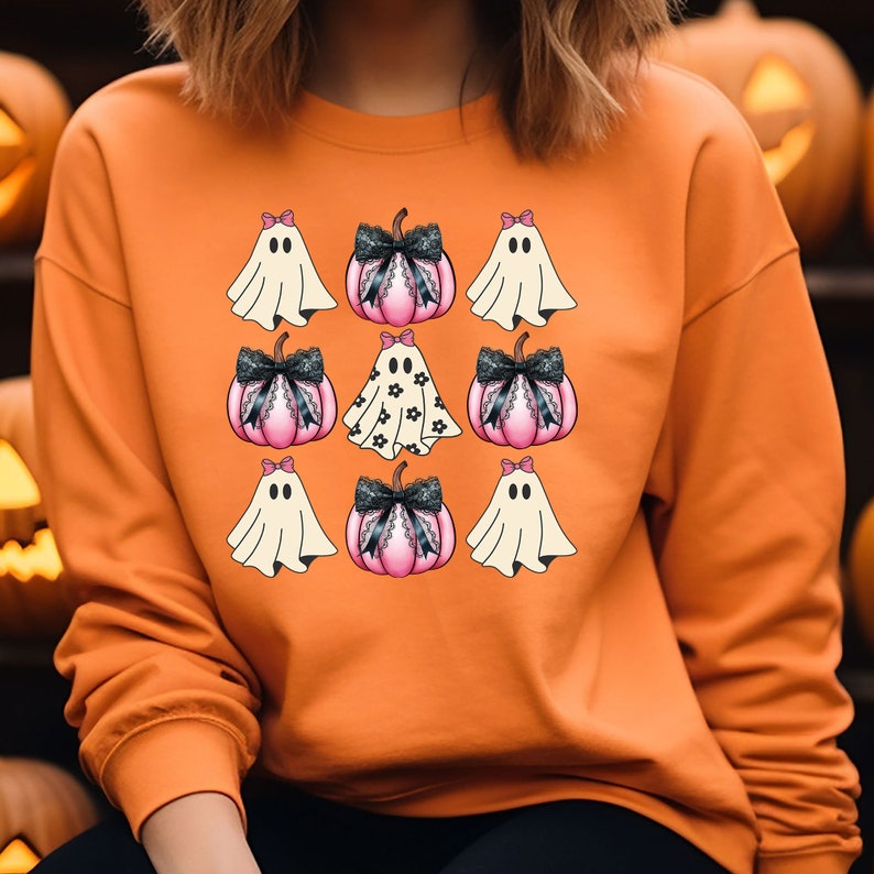May include: Orange sweatshirt featuring a grid of Halloween-themed graphics. The design includes cartoon ghosts with bows and pink pumpkins with black lace bows. The central pumpkin has a floral pattern. The overall theme is festive and seasonal.