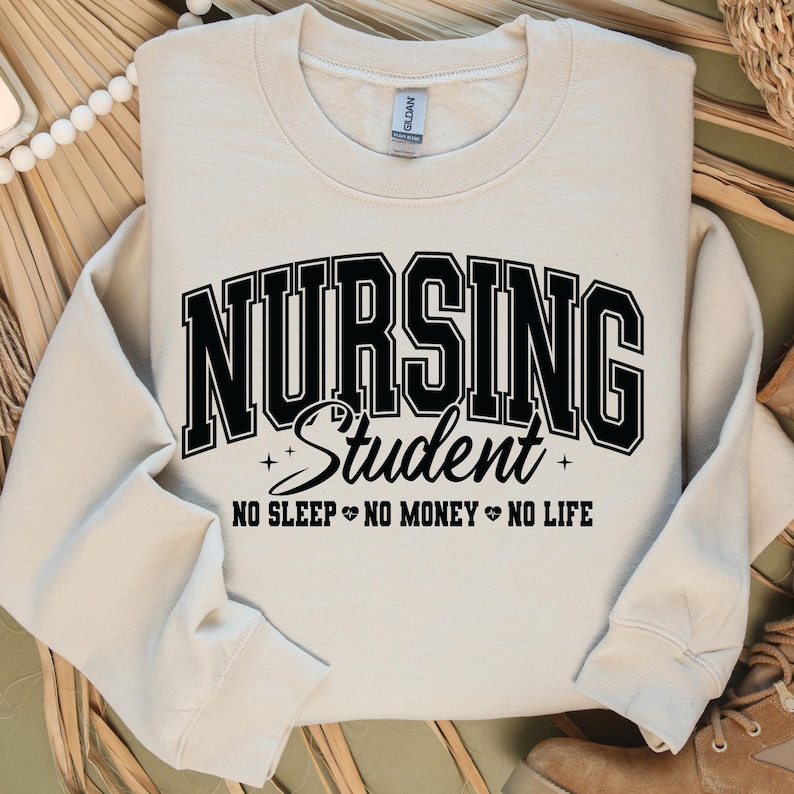 Op de afbeelding: Cr&egrave;mekleurige sweatshirt met de woorden "NURSING Student" in vette zwarte letters. Daaronder staat de tekst "NO SLEEP NO MONEY NO LIFE". De sweatshirt heeft een ronde hals en lange mouwen.