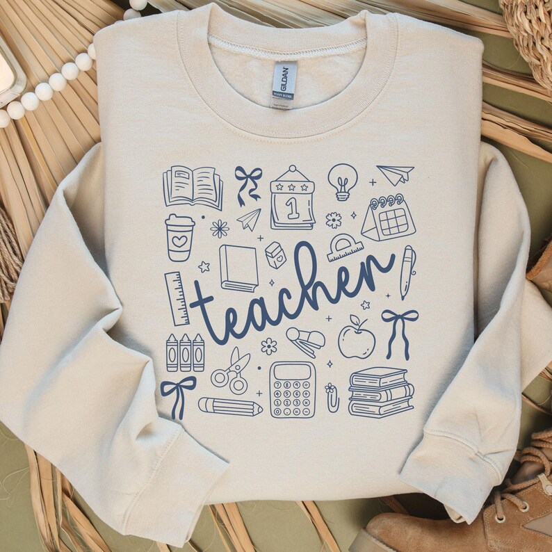 May include: A cream-colored sweatshirt with a blue graphic design featuring school-related icons and the word "teacher". The design includes books, pencils, an apple, and a calculator.