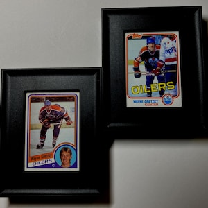 May include: Two framed hockey cards featuring Wayne Gretzky in an Edmonton Oilers uniform. The cards are in black frames, showcasing the player in action and a portrait. The cards have the text "OILERS" and "WAYNE GRETZKY CENTER".
