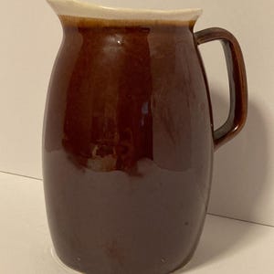 May include: A ceramic pitcher with a glossy dark brown glaze covering the body and a contrasting cream-colored rim. The vessel features a rounded, bulbous shape, a wide pouring spout, and a sturdy, curved handle attached to the side.