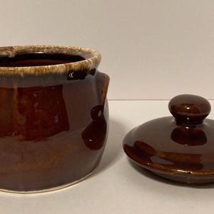 May include: A round, dark brown ceramic bean pot with two side handles and a matching lid. The pot features a glossy, smooth finish with a lighter, mottled beige glaze along the upper rim and the top edge of the lid, which has a small knob handle.