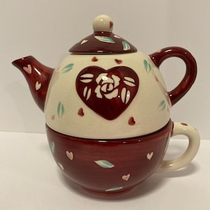 May include: A ceramic tea for one set featuring a cream-colored teapot with a dark red lid, handle, and spout. The teapot has a large dark red heart containing a white rose design, accented by small red hearts and light green leaves.