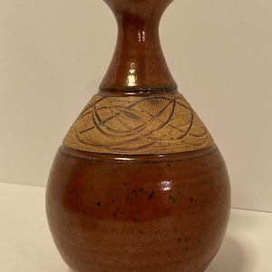 May include: A ceramic vase with a bulbous base and a slender neck. The piece features a glossy, deep reddish-brown glaze on the neck and lower body, contrasted by a textured, matte tan band around the shoulder with incised swirling line patterns.