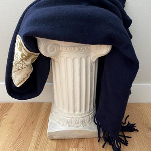 May include: A navy blue wool blanket with fringe detail draped over a white, classical-style pedestal. The blanket has a decorative patch with a gold and cream-coloured design on one corner. The blanket is displayed on a light wood floor.