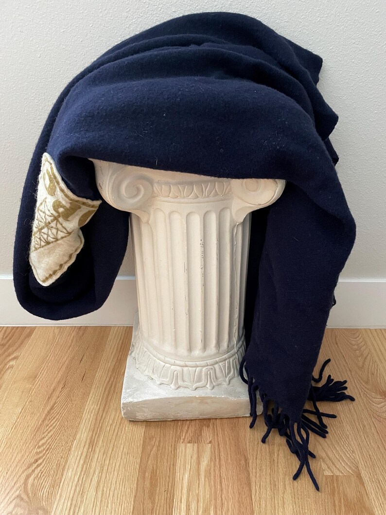 May include: A navy blue wool blanket with fringe detail draped over a white, classical-style pedestal. The blanket has a decorative patch with a gold and cream-coloured design on one corner. The blanket is displayed on a light wood floor.