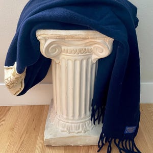 May include: A navy blue scarf with fringing draped over a white decorative column. The scarf has a cream border with gold embroidery. The column is made of a stone-like material and has a classical design.