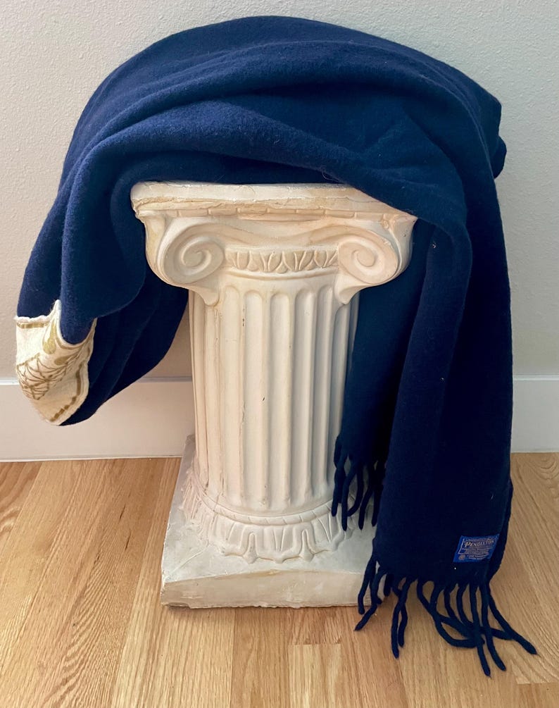 May include: A navy blue scarf with fringing draped over a white decorative column. The scarf has a cream border with gold embroidery. The column is made of a stone-like material and has a classical design.