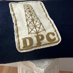 May include: A navy blue blanket with a rectangular patch featuring a brown oil derrick graphic and the letters "DPC". The blanket is folded and resting on a white surface.