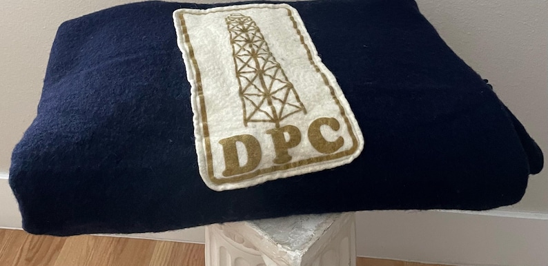 May include: A navy blue blanket with a rectangular patch featuring a brown oil derrick graphic and the letters "DPC". The blanket is folded and resting on a white surface.