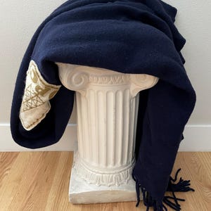 May include: A navy blue blanket with a decorative patch and fringe detail. The blanket is draped over a white, classical-style pedestal. The patch features a cream-coloured background with gold embroidery.