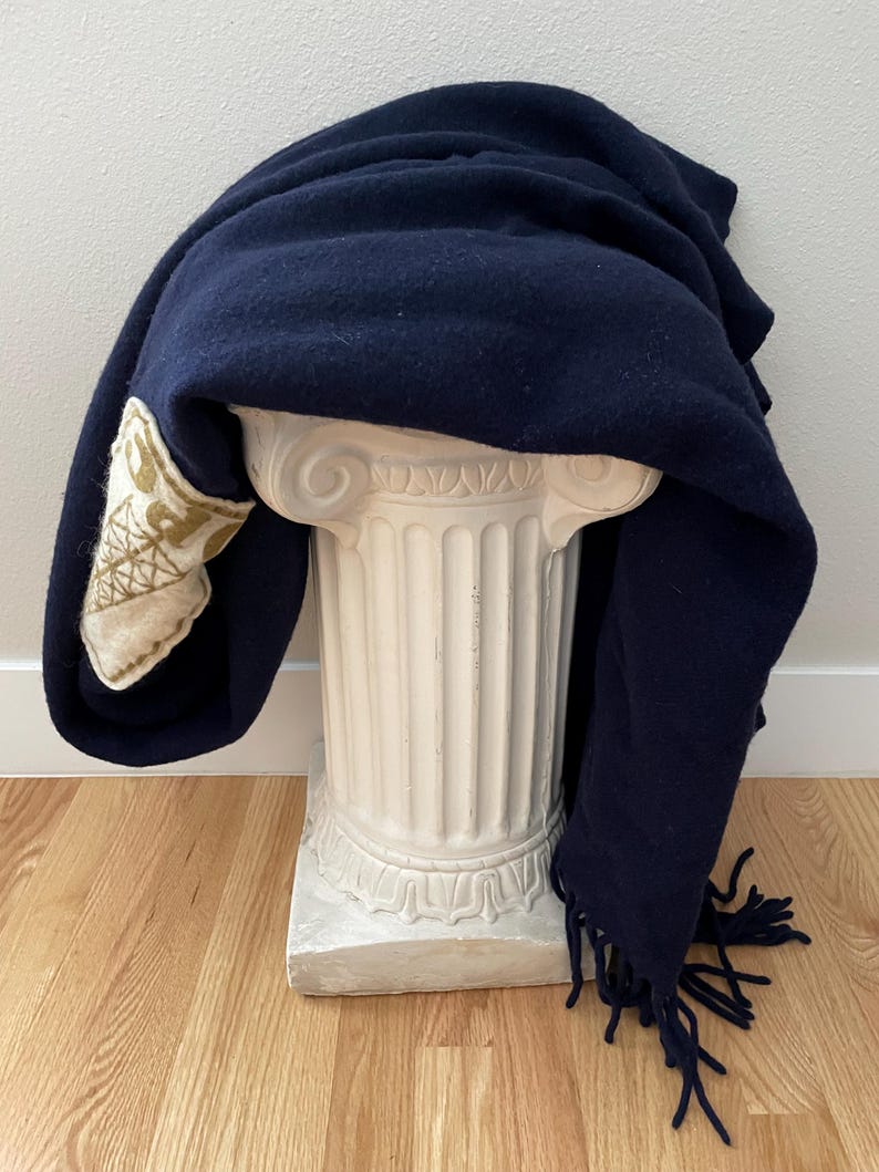 May include: A navy blue blanket with a decorative patch and fringe detail. The blanket is draped over a white, classical-style pedestal. The patch features a cream-coloured background with gold embroidery.