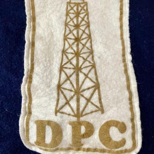 May include: A white felt patch featuring a brown oil derrick design and the letters "DPC" at the base. The patch has a brown border and is set against a dark blue background.