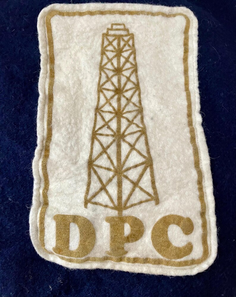 May include: A white felt patch featuring a brown oil derrick design and the letters "DPC" at the base. The patch has a brown border and is set against a dark blue background.