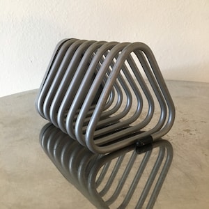 May include: A silver-colored metal rack with a series of triangular loops. The loops are arranged in a row, creating a geometric design. The rack is resting on a reflective surface, with its mirrored image visible below.