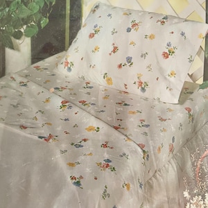 May include: A white floral patterned bed set featuring a pillow and sheet. The pattern includes small, colorful flowers in shades of red, yellow, and blue. The bedding is made of a soft, possibly cotton, material.