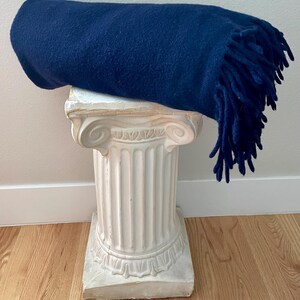 May include: A rolled navy blue blanket with fringing rests on a white, classical-style pedestal. The blanket's texture is visible, and the fringing hangs down. The pedestal has a detailed, ornate design.