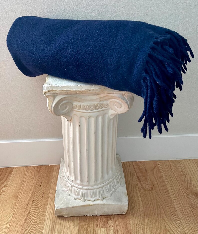 May include: A rolled navy blue blanket with fringing rests on a white, classical-style pedestal. The blanket's texture is visible, and the fringing hangs down. The pedestal has a detailed, ornate design.