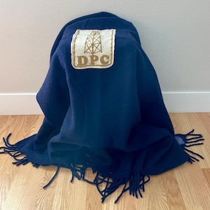 May include: A navy blue blanket with a fringed edge. A rectangular patch with the text "DPC" and an illustration of an oil derrick is sewn onto the blanket. The blanket is folded and draped.