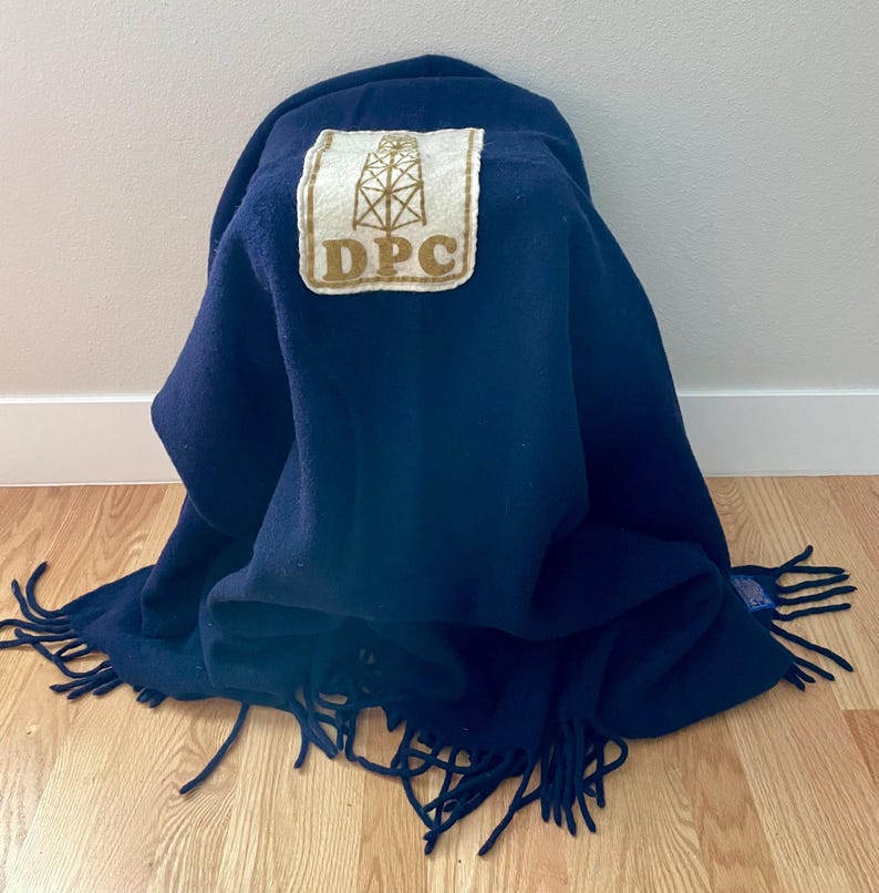 May include: A navy blue blanket with a fringed edge. A rectangular patch with the text "DPC" and an illustration of an oil derrick is sewn onto the blanket. The blanket is folded and draped.
