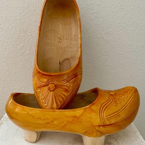 May include: Two wooden clogs, one resting atop the other. The clogs are a warm, golden-yellow color with carved floral designs on the front. The lower clog has three small, off-white feet. The clogs appear to be handmade.