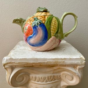 May include: A ceramic teapot with a vegetable design. The teapot features a tomato, eggplant, corn, and other vegetables. The handle and spout are green, and the lid is topped with a leaf. The teapot is sitting on a white, decorative pedestal.