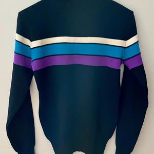 May include: A black sweater with horizontal stripes in white, blue, and purple. The sweater has long sleeves and a ribbed hem and cuffs. The sweater is hanging on a wooden hanger against a white wall.