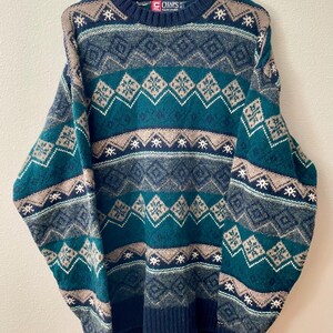 May include: A vintage Chaps sweater in teal, navy, and tan with a geometric pattern. The crew neck sweater features a diamond and snowflake design. The sweater is hanging on a wooden hanger.