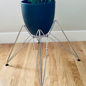 May include: A matte black cylindrical planter held by a chrome-finished metal wire stand with four angled legs. The planter contains a green leafy plant. The stand features thin, intersecting metal rods and small black protective feet at the base.