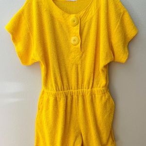 May include: Bright yellow terry cloth romper with short sleeves and shorts. The romper features a round neckline, two large buttons, and an elastic waist. The garment is hanging on a wooden hanger.