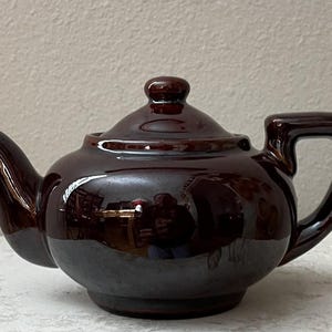 May include: A glossy, dark brown ceramic teapot with a rounded body, a curved spout, and a square handle. The lid has a small knob. The teapot sits on a white surface. A classic kitchenware item.