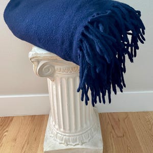 May include: A navy blue blanket with fringing is draped over a white decorative column. The blanket has a soft, textured appearance. The column has a classic, architectural design, and is placed on a wooden floor.
