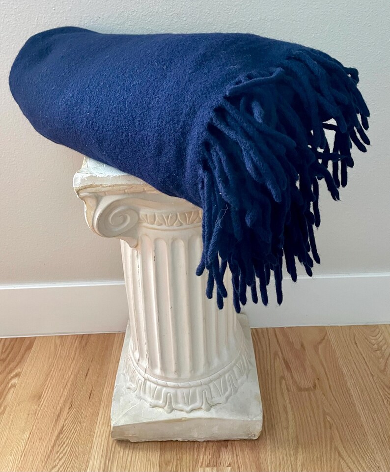 May include: A navy blue blanket with fringing is draped over a white decorative column. The blanket has a soft, textured appearance. The column has a classic, architectural design, and is placed on a wooden floor.