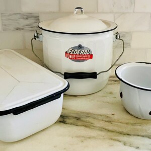 May include: Three white enamelware kitchen items: a rectangular covered dish with a black rim, a round covered pot with a black handle and a metal handle, and a small round bowl with a black rim and handle. The pot has a red and white label that reads "Federal Vogue" with the words "enameledware" and "made in the USA" in smaller font.