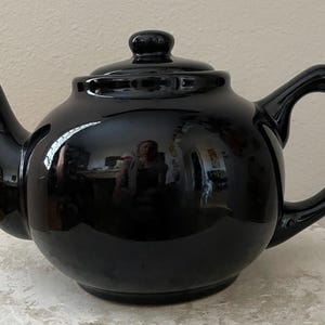 May include: A glossy black ceramic teapot with a rounded body, spout, and handle. The lid has a small knob. The teapot is sitting on a textured surface. The teapot reflects the surrounding environment.