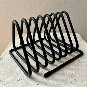 May include: A black metal desk organizer with a series of angled, curved supports. The organizer has a rectangular base and a rod extending from the side. The design is minimalist and modern.