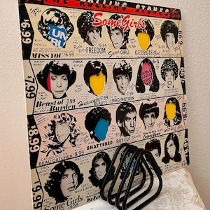 May include: A vintage record album cover for The Rolling Stones album "Some Girls". The cover features a collage of different hairstyles and hair colors, with each style labeled with a name and price. The album title is printed in large, bold letters at the top of the cover.