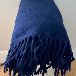 May include: A navy blue blanket with a textured surface and long fringe. The blanket is draped over a white decorative pedestal. The blanket is made of a soft material.