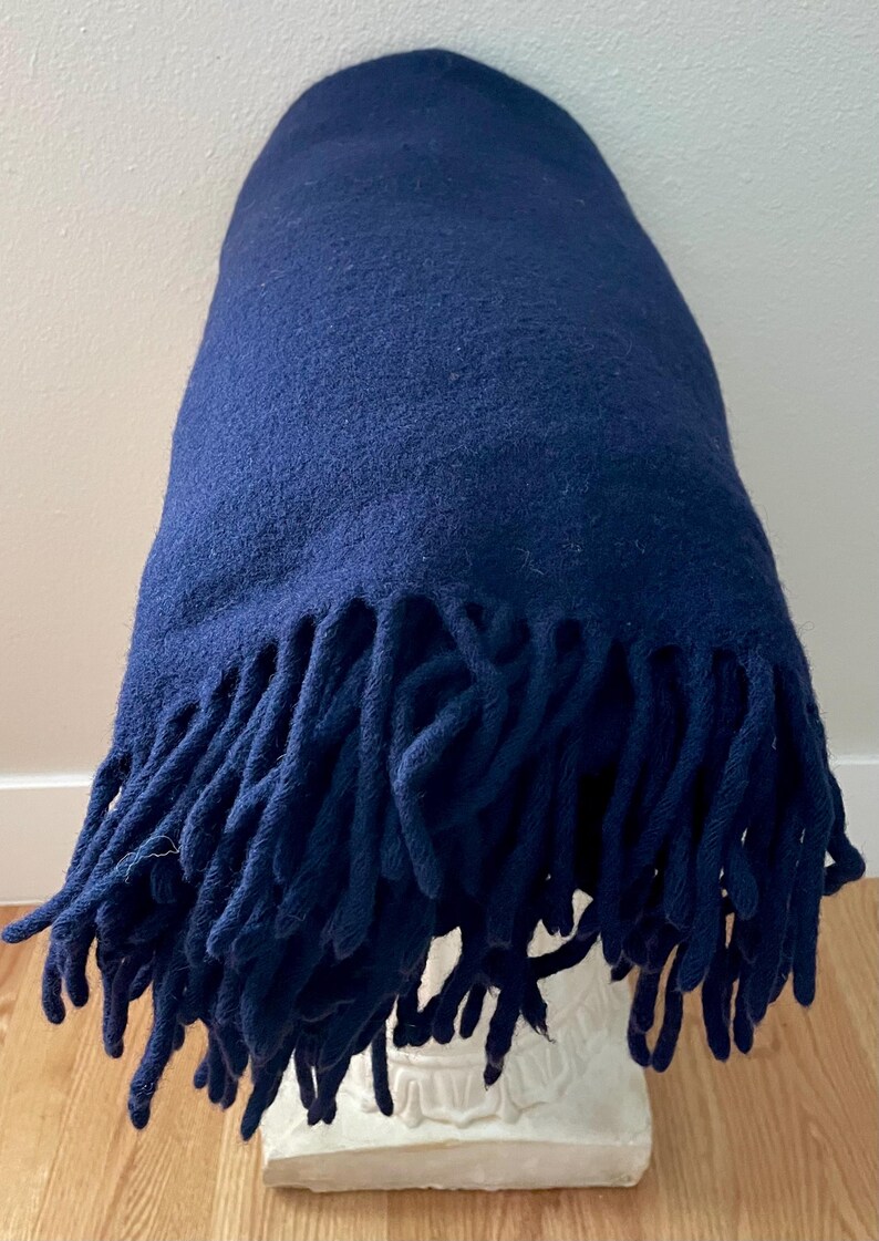 May include: A navy blue blanket with a textured surface and long fringe. The blanket is draped over a white decorative pedestal. The blanket is made of a soft material.