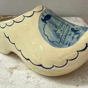 May include: A ceramic Dutch clog planter in a cream color with blue and brown accents. The clog features a blue illustration of a ship and wavy blue trim. The interior is brown.