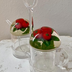 May include: Clear plastic salt and pepper shakers with a handle. Each shaker has a clear dome lid with a design of red tomatoes and green leaves. The set includes two small spoons.