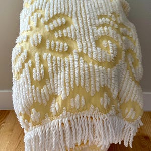 May include: A yellow and white tufted throw blanket with a fringe trim. The blanket features a repeating pattern of white tufts on a yellow background. The fringe is a thick, white rope-like material.