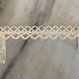 May include: A white, decorative wall shelf with a delicate, openwork design. The shelf features a scalloped edge with a repeating floral pattern. Two curved brackets support the shelf, creating a floating appearance. The shelf is made of metal.